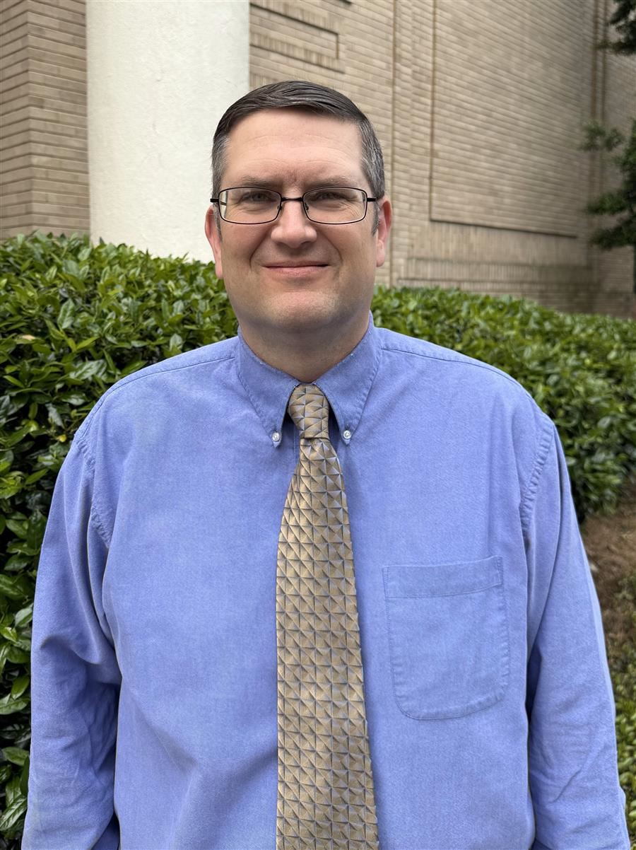 Jason Roop wears a blue shirt & beige patterned tie standing outdoors in front of a brick wall.